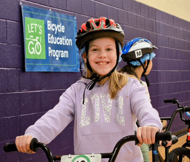 A child wearing a bike helmet smiles while holding handlebars during an indoor Let’s Go bicycle education session