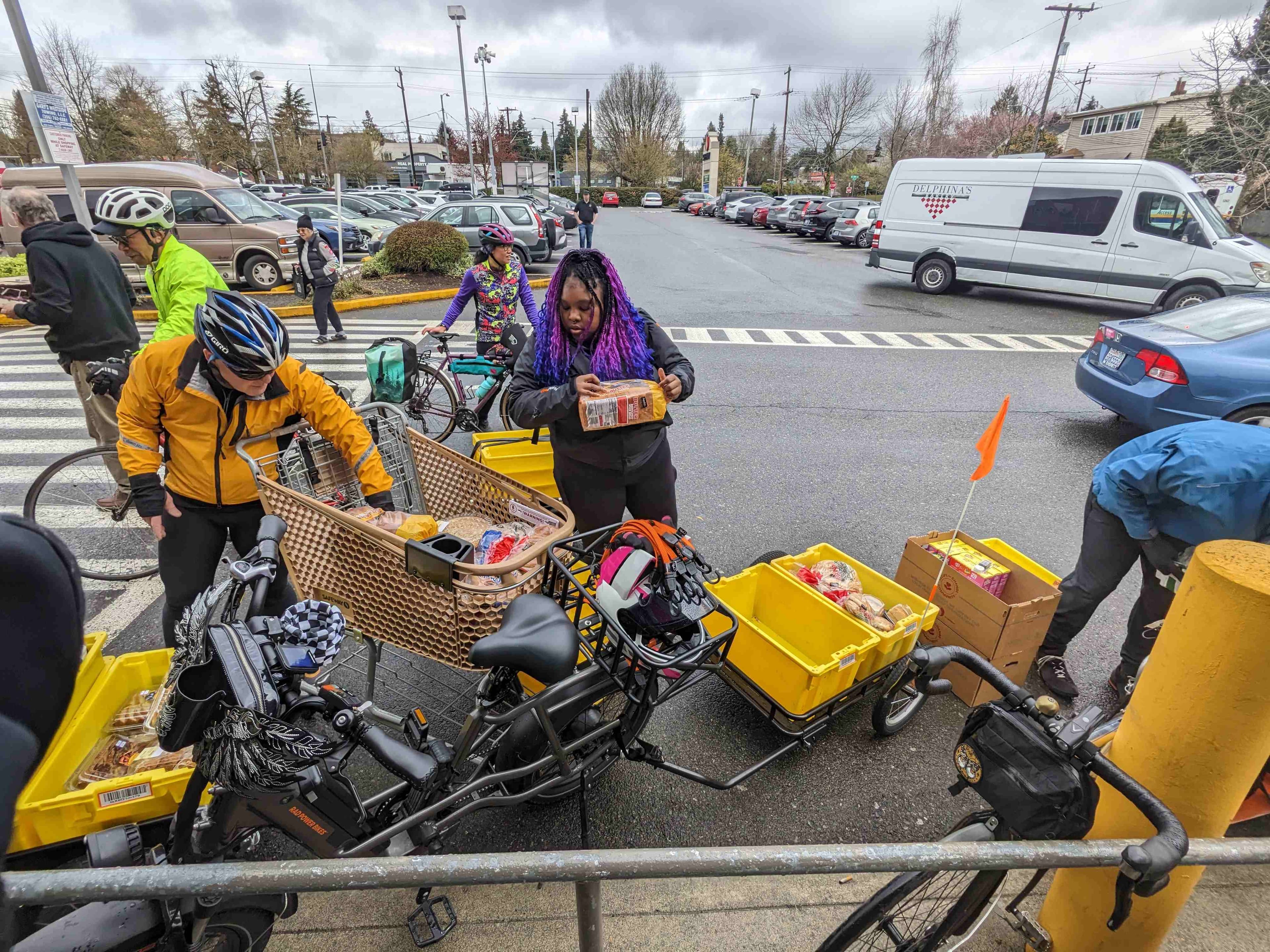 Community members with cargo bikes distributing supplies in parking lot, including person with purple braids examining items from yellow cargo containers.